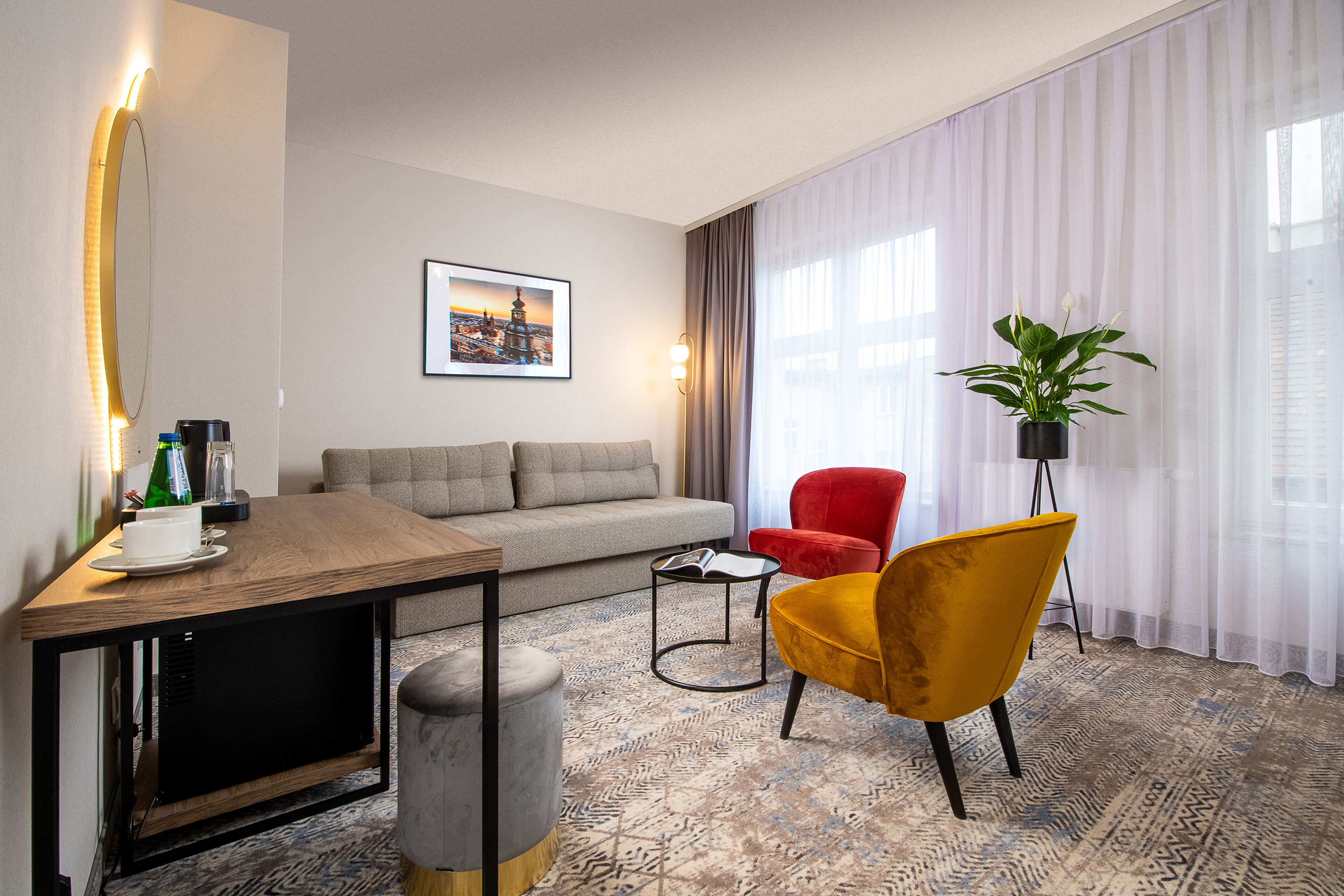 A modern hotel room with a sofa, a red armchair, a small table with a cup and saucer, a floor lamp, and a large window with sheer curtains.