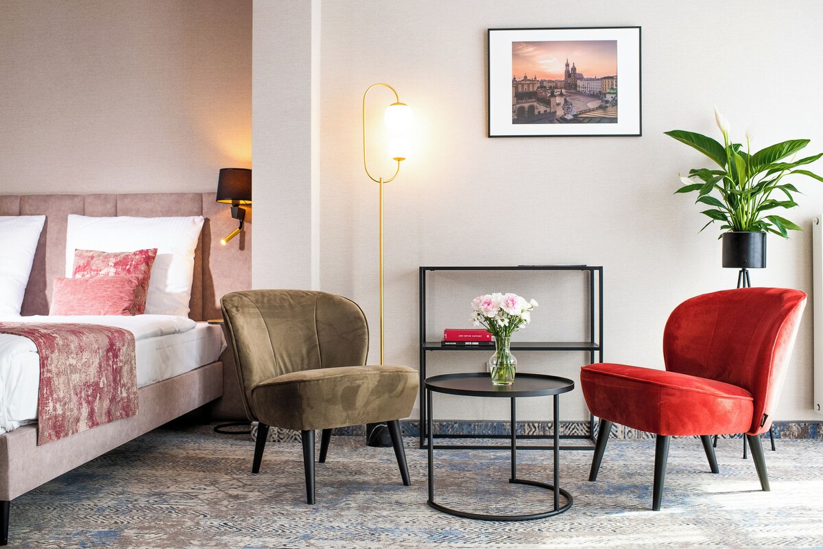 A hotel room with a bed, two chairs, a small table with a vase, a floor lamp, a framed picture on the wall, and a potted plant.