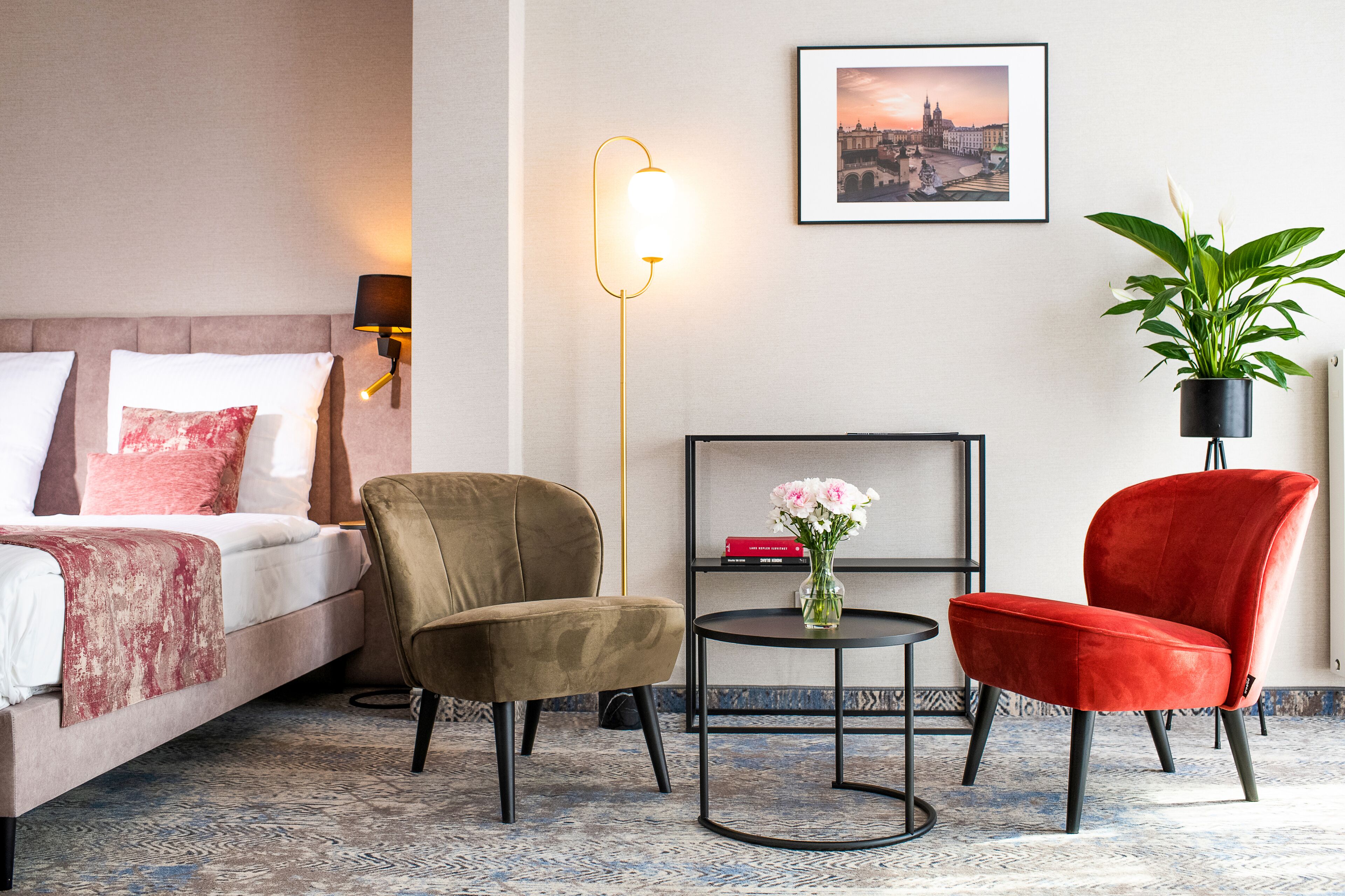 Une chambre d’hôtel avec un lit, deux chaises, une petite table sur laquelle se trouve un vase, une lampe sur pied, un tableau encadré au mur et une plante en pot.
