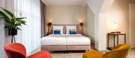 A hotel room with a large bed, two bedside tables with lamps, a small side table with a lamp, a potted plant, and two chairs.