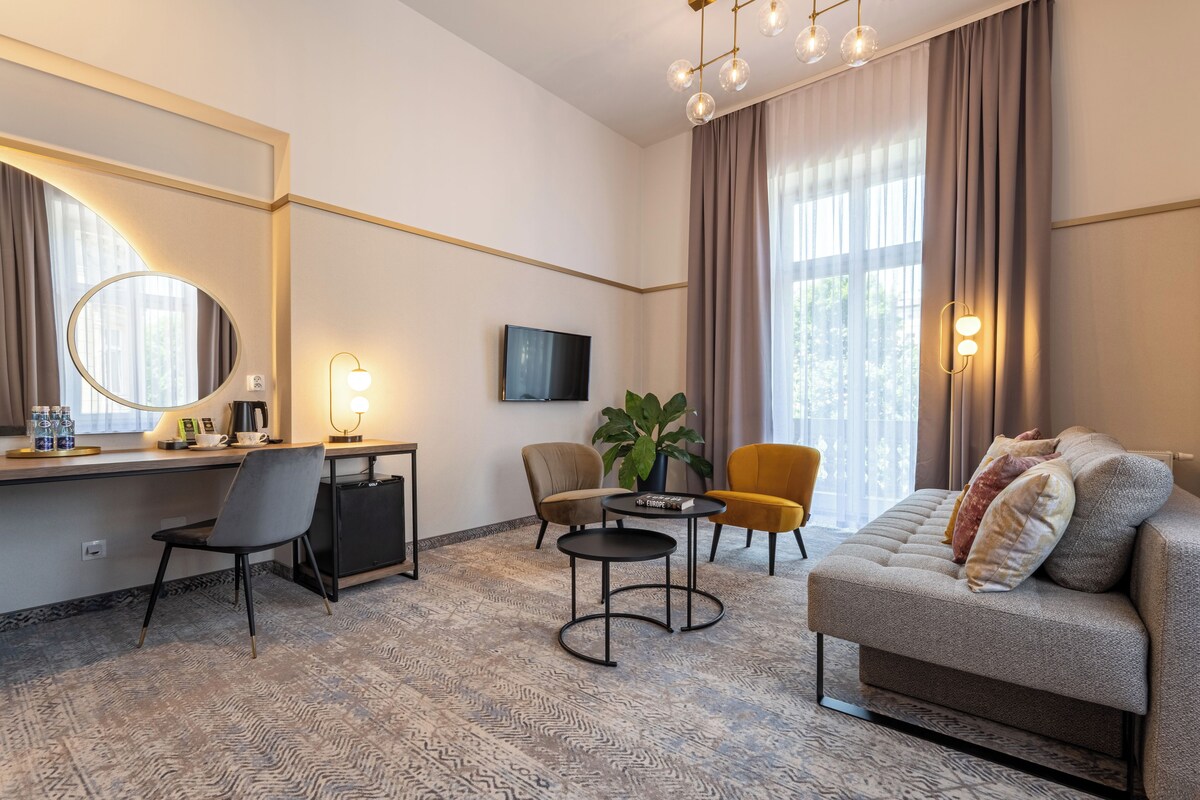 A modern hotel room with a sofa, a desk with a chair, a small table, a TV, and a large window with curtains.