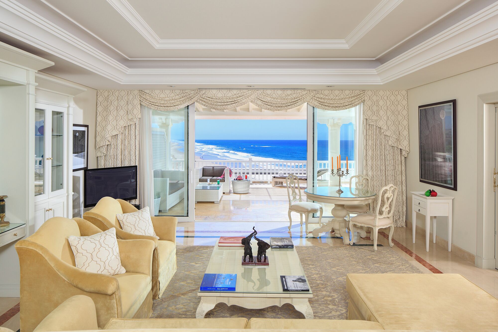 Amplia sala de estar con un gran ventanal con vista a la playa, un televisor de pantalla plana y un comedor con mesa y sillas.