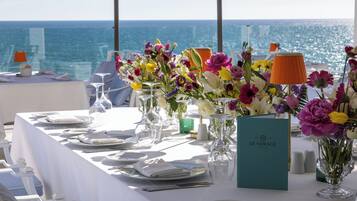 Ein Tisch ist für eine Mahlzeit gedeckt, mit Blick auf das Meer, inklusive Speisekarte, Gläsern und floralen Tischdekorationen.