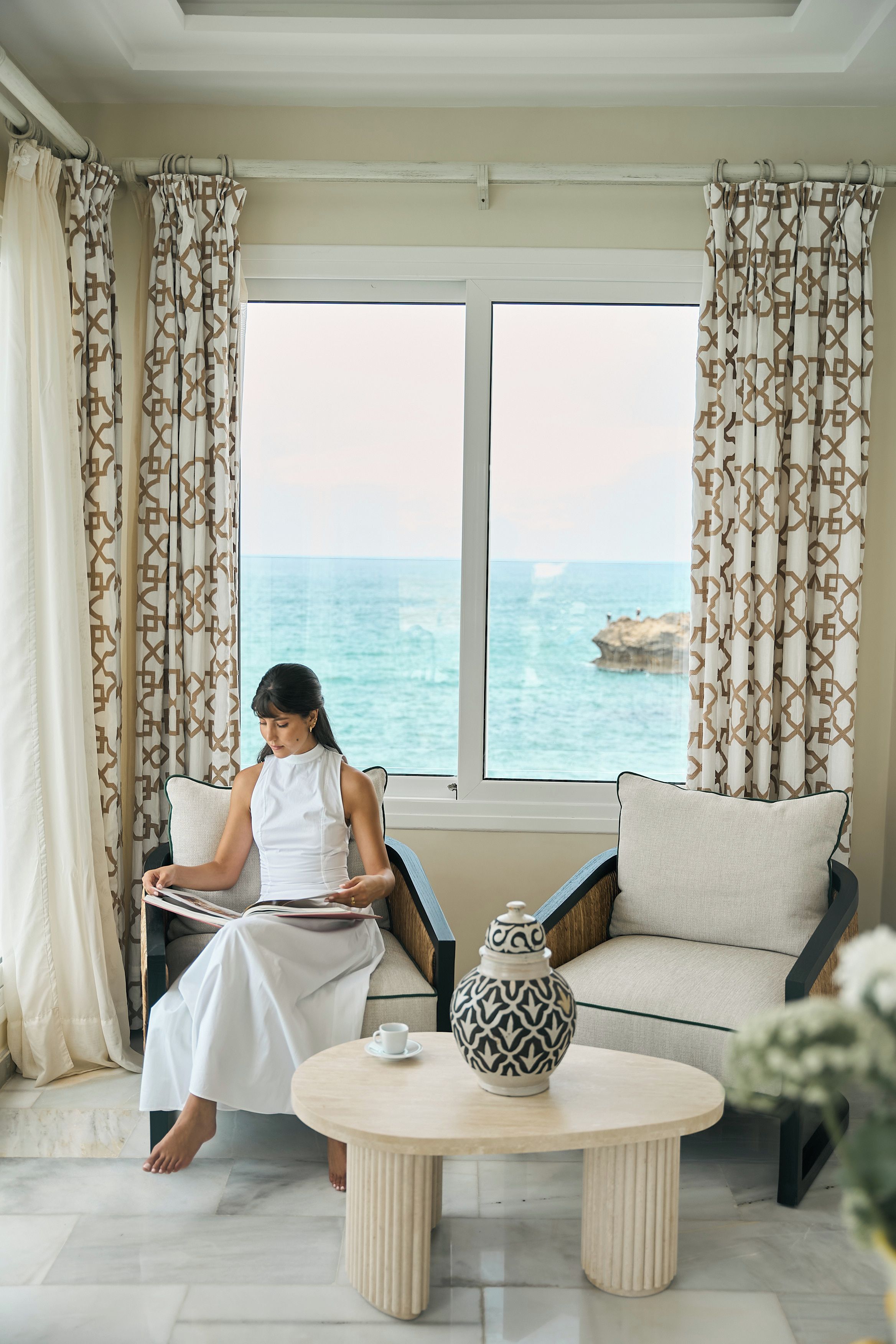 Une pièce dotée d’une grande fenêtre donnant sur la mer, d’un canapé, d’une table basse agrémentée d’un vase décoratif, et d’une personne lisant un livre.