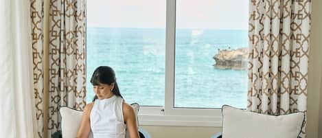 Una habitación con un ventanal grande con vistas al mar, un sofá, una mesa de centro con un jarrón decorativo y una persona leyendo un libro.