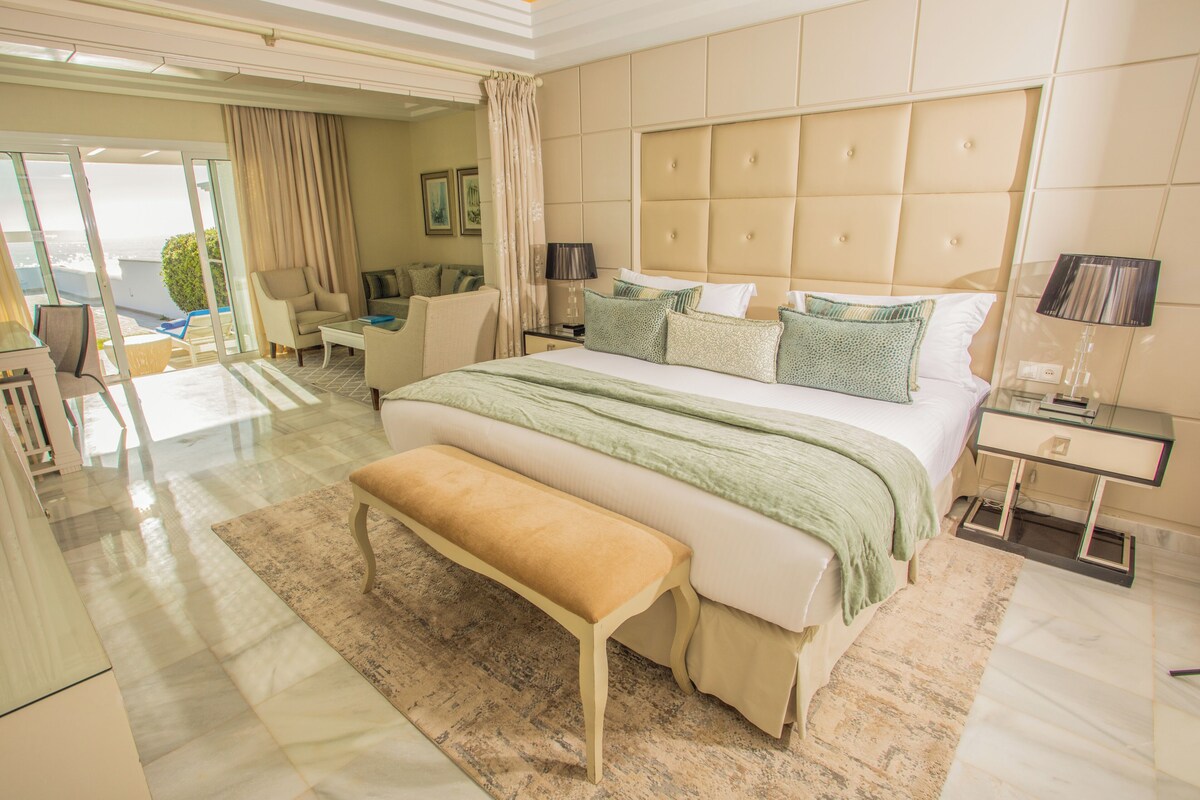 A modern bedroom with a large bed, bedside tables, a bench, and a view of the outdoors.