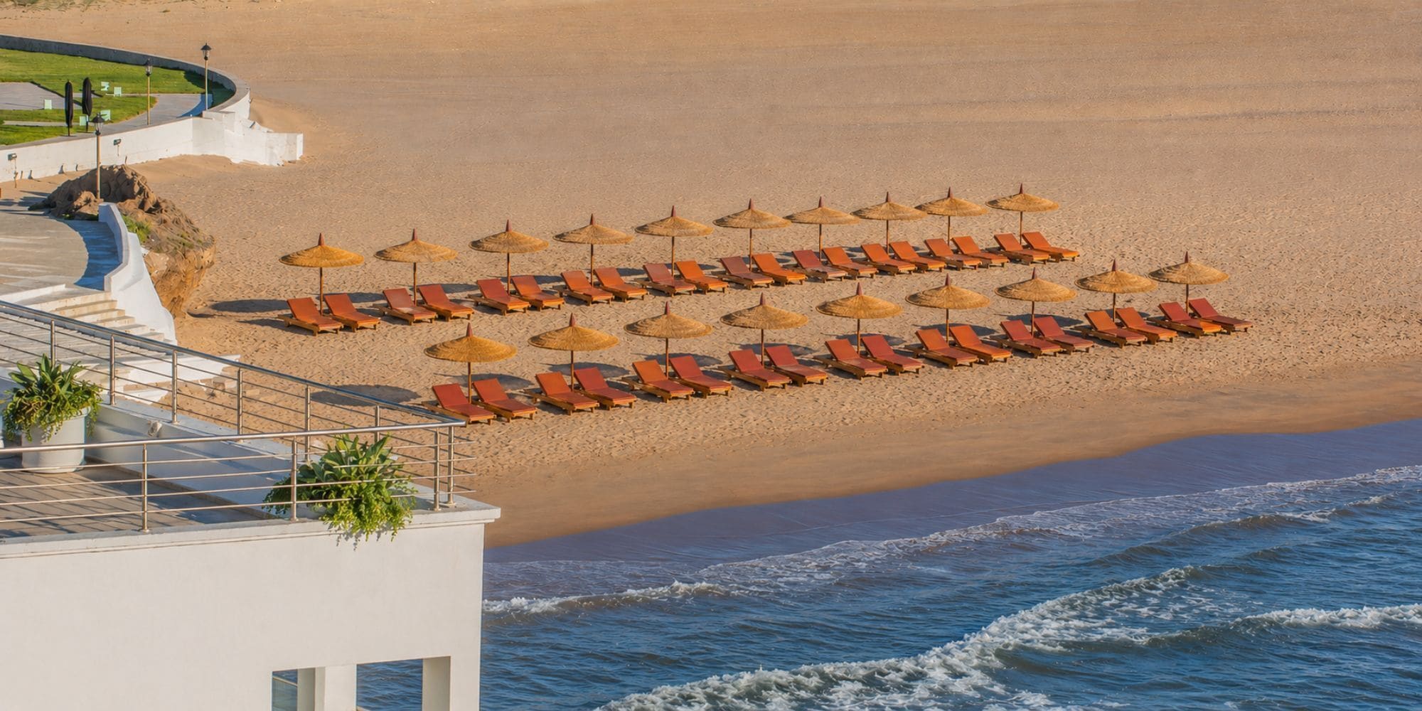 Un hôtel situé en bord de mer, avec des rangées de chaises longues et des parasols en chaume, un espace piscine et une vue sur l’océan.