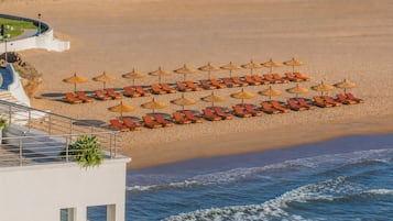 A beachside hotel with rows of sun loungers and thatched umbrellas, a pool area, and a view of the ocean.