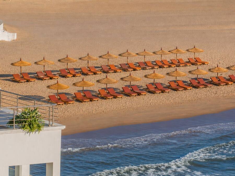 Un hotel frente a la playa con filas de tumbonas y sombrillas de paja, un área de piscina y vistas al océano.