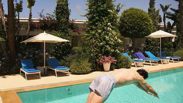 Une personne plonge dans une piscine à l’eau claire et bleue, avec des chaises longues et des palmiers en arrière-plan.