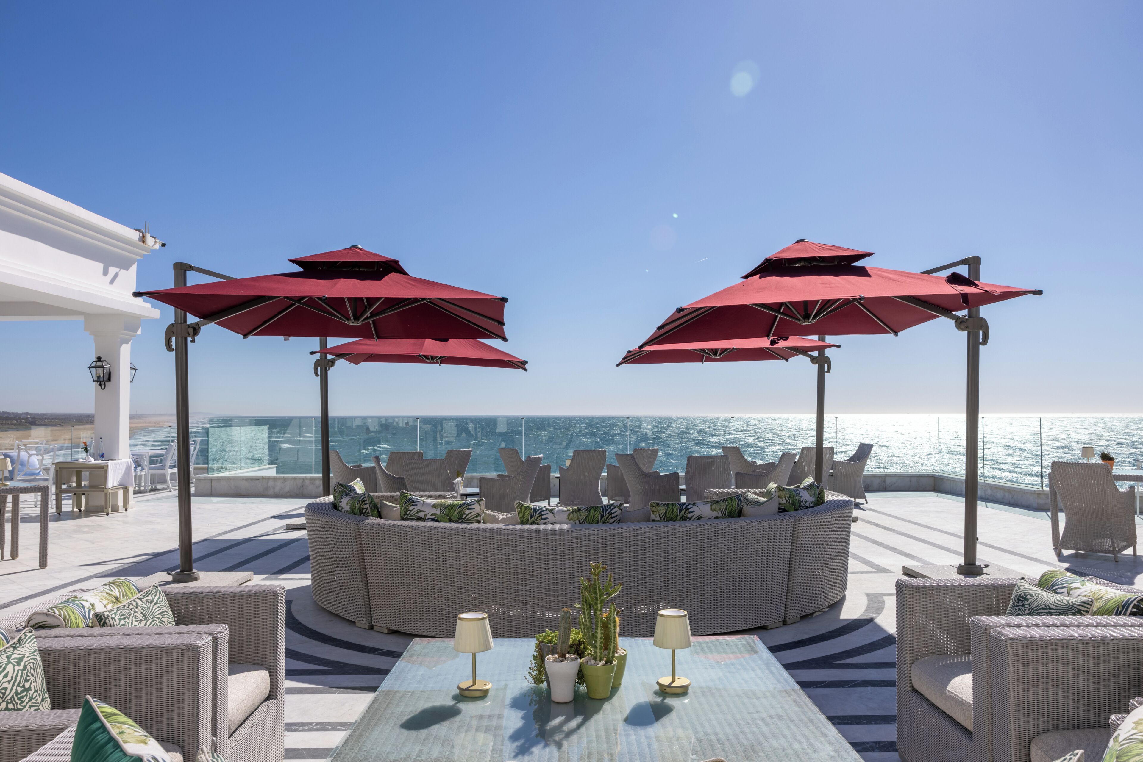 Une terrasse en bord de mer, avec des meubles en osier, des parasols rouges et une vue sur l’océan.