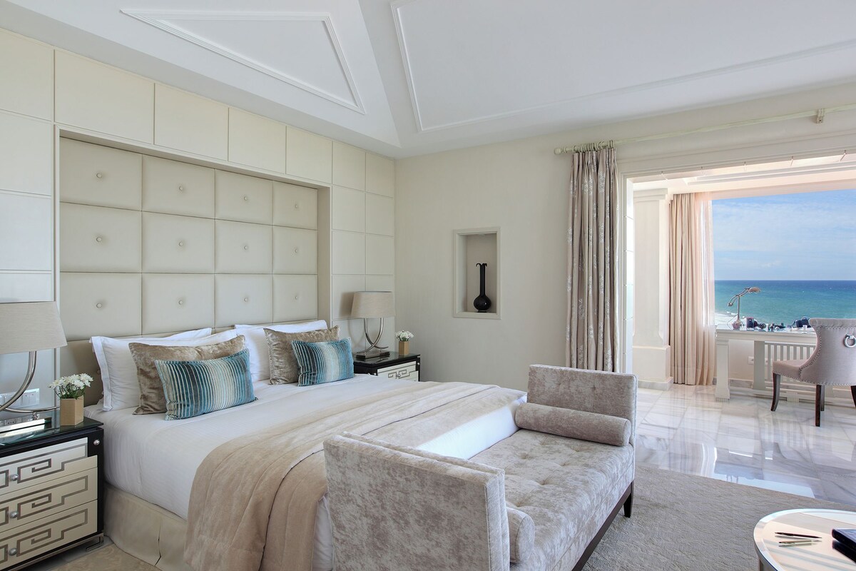 Habitación de hotel con una cama grande, zona de estar, vista al mar y un balcón.