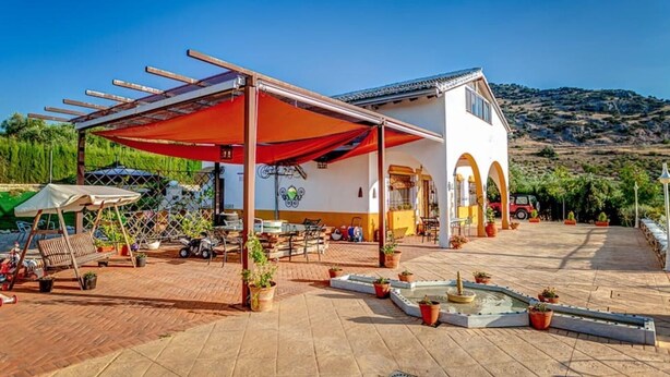 Ein weißes Gebäude mit einer überdachten Terrasse, Außenbestuhlung und einem kleinen Brunnen.
