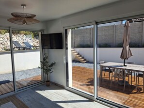 A modern room with a large sliding glass door leading to an outdoor patio with a wooden deck, a table with chairs, a potted plant, and a TV mounted on the wall.