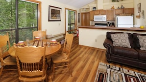 A dining area with a wooden table and chairs, a kitchen with wooden cabinets and appliances, and a living room with a brown leather couch and patterned cushions. - The Bird Hallar - Cozy, quiet cabin in The Glades! (Gatlinburg)