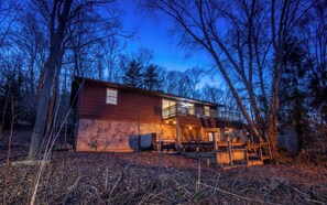 A cabin with a balcony, surrounded by trees, and a deck area with outdoor seating.