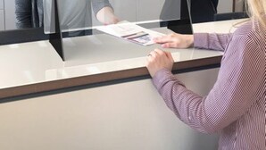 Un mostrador de recepción con una mampara de vidrio, una computadora y un folleto en la barra.