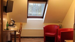Una habitación pequeña y acogedora con techo inclinado, ventana con vista a árboles, una silla roja, una mesa de madera con una silla, y un televisor montado en la pared.