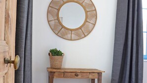 Una habitación con puerta de madera, un espejo redondo en la pared, una pequeña mesa de madera con una cesta de plantas verdes y cortinas grises.