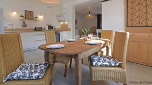 Un comedor con una mesa de madera dispuesta para cuatro personas, rodeado de sillas de mimbre con cojines estampados. Hay lámparas colgantes sobre la mesa, y se ve una zona de cocina al fondo.