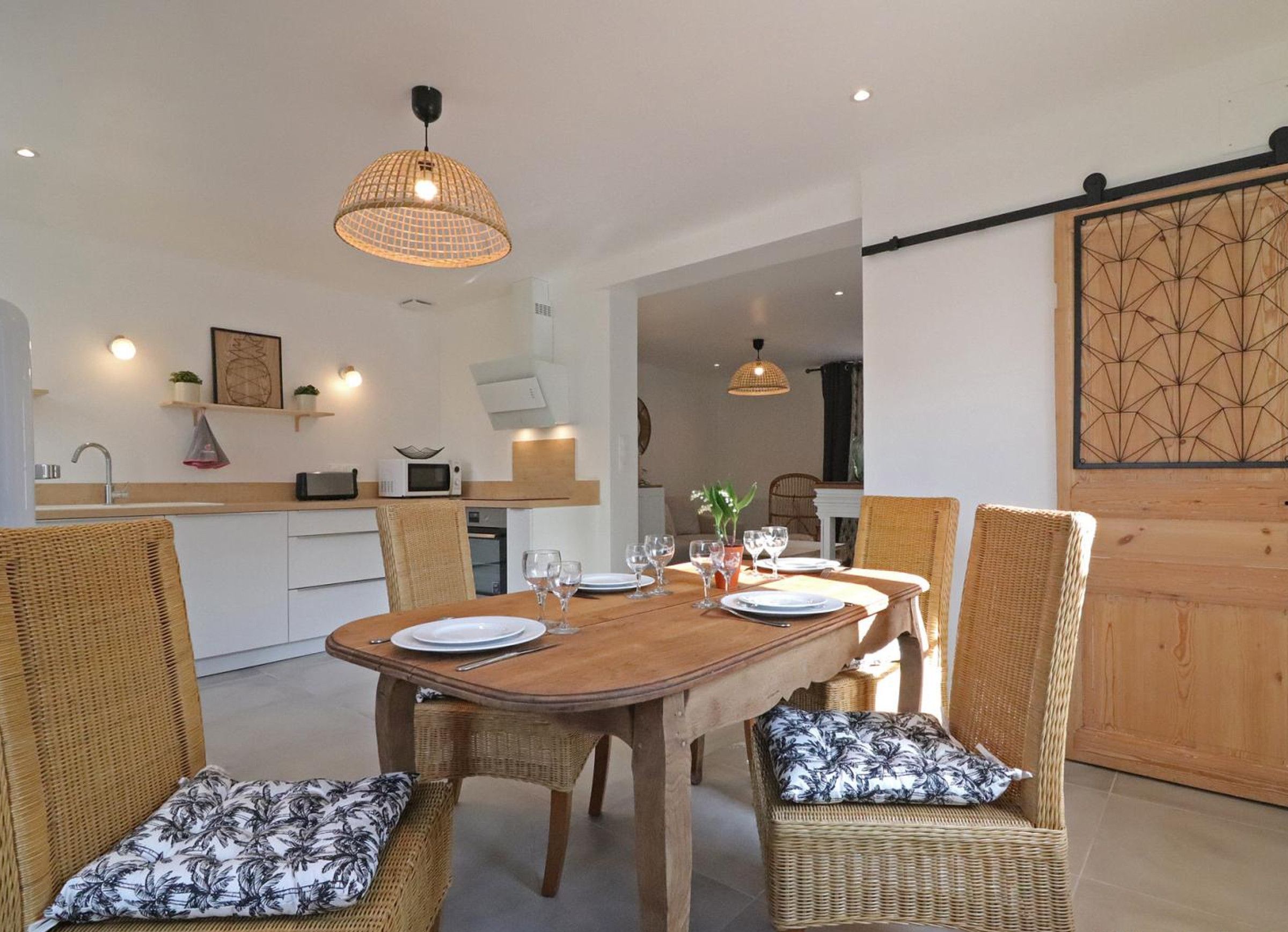 Un comedor con una mesa de madera dispuesta para cuatro personas, rodeado de sillas de mimbre con cojines estampados. Hay lámparas colgantes sobre la mesa, y se ve una zona de cocina al fondo.