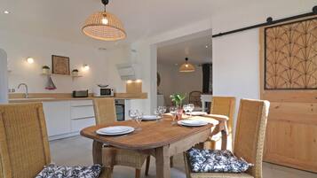 Un comedor con una mesa de madera dispuesta para cuatro personas, rodeado de sillas de mimbre con cojines estampados. Hay lámparas colgantes sobre la mesa, y se ve una zona de cocina al fondo.