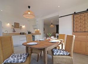 Ein Essbereich mit einem Holztisch, gedeckt für vier Personen, umgeben von Korbstühlen mit gemusterten Kissen. Über dem Tisch hängen Pendelleuchten, und im Hintergrund ist eine Küche erkennbar.