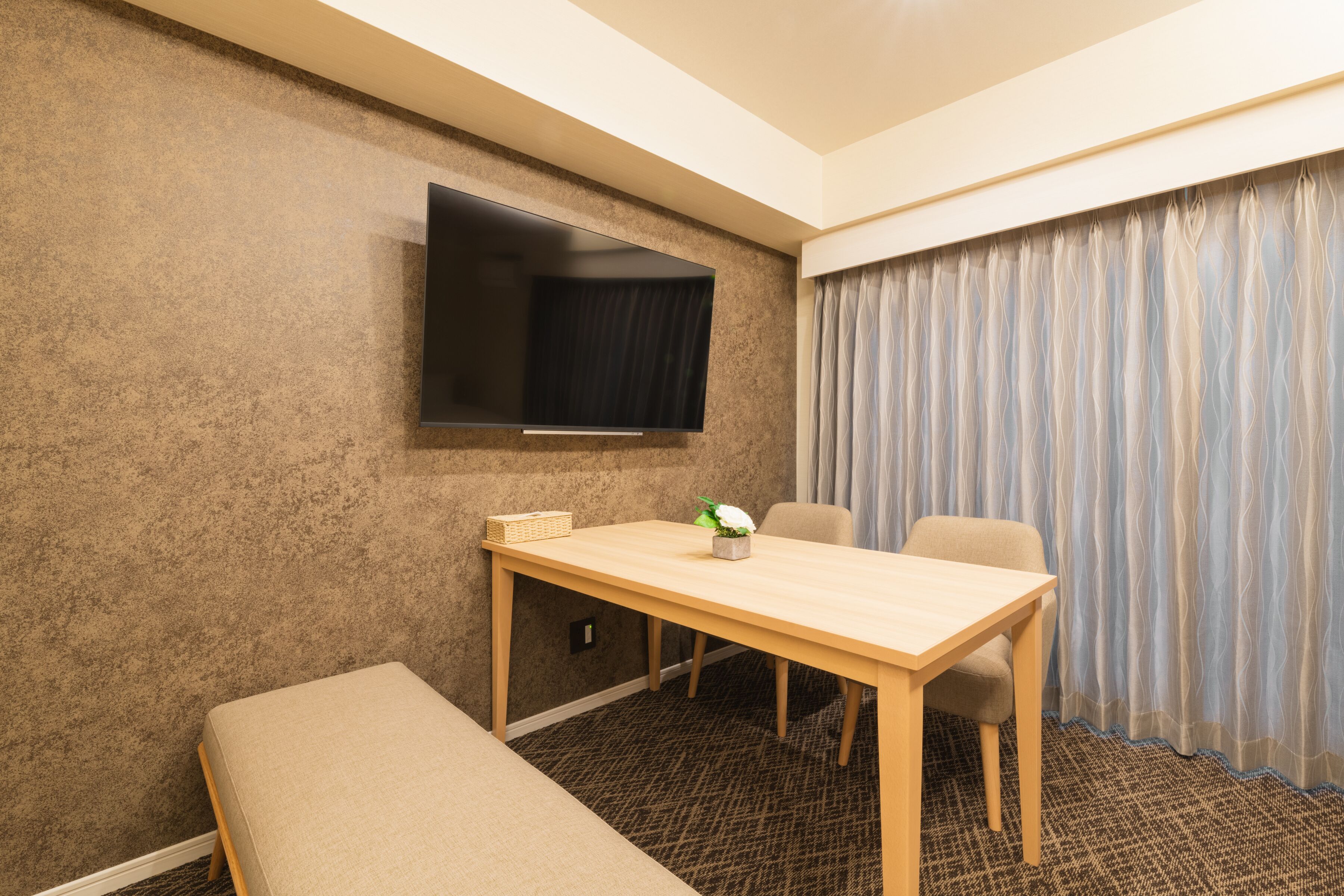 A hotel room with a wooden table, two chairs, a bench, a flat-screen TV mounted on the wall, and a patterned carpet.