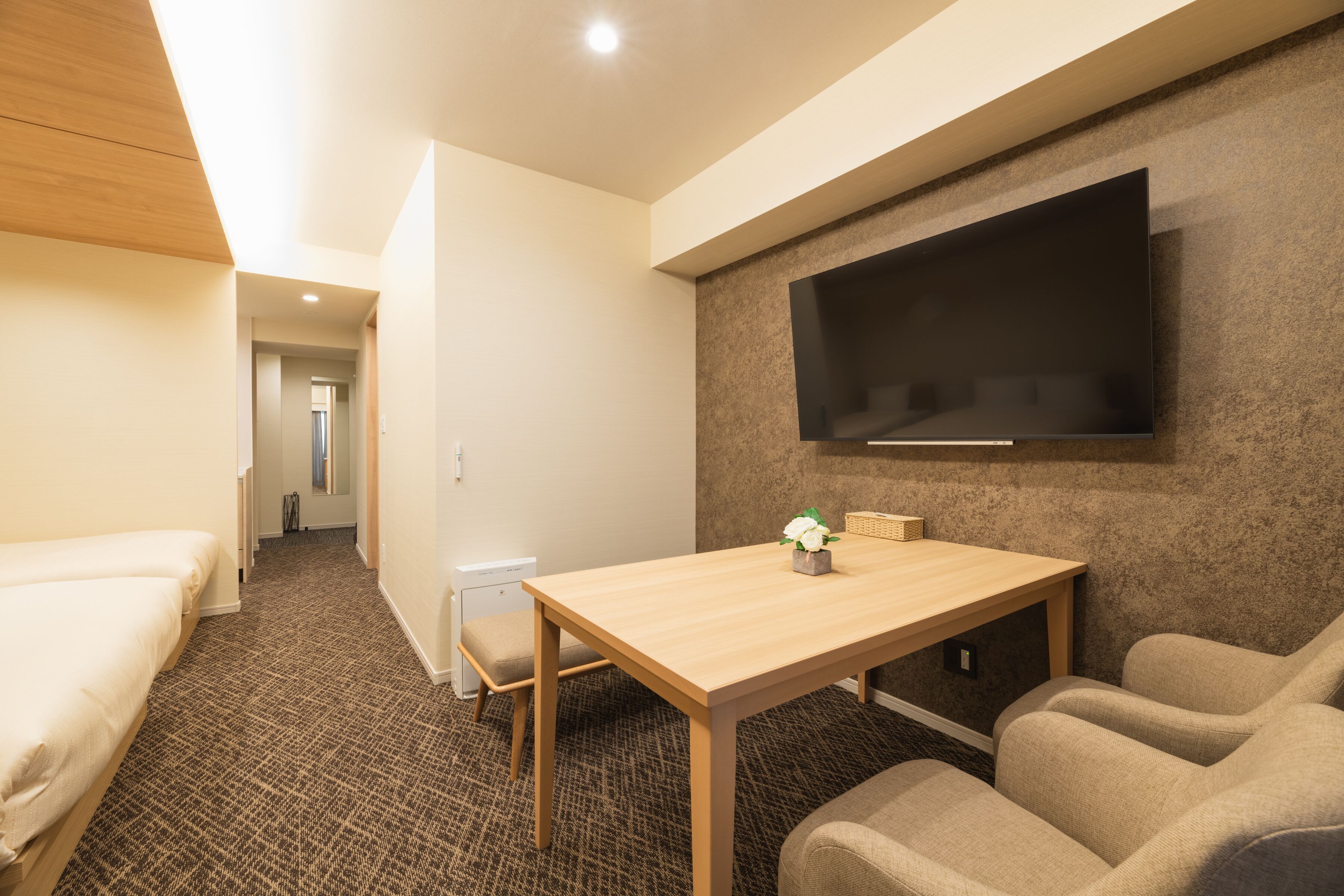 A hotel room with a bed, a desk, a chair, a TV mounted on the wall, and a carpeted floor.