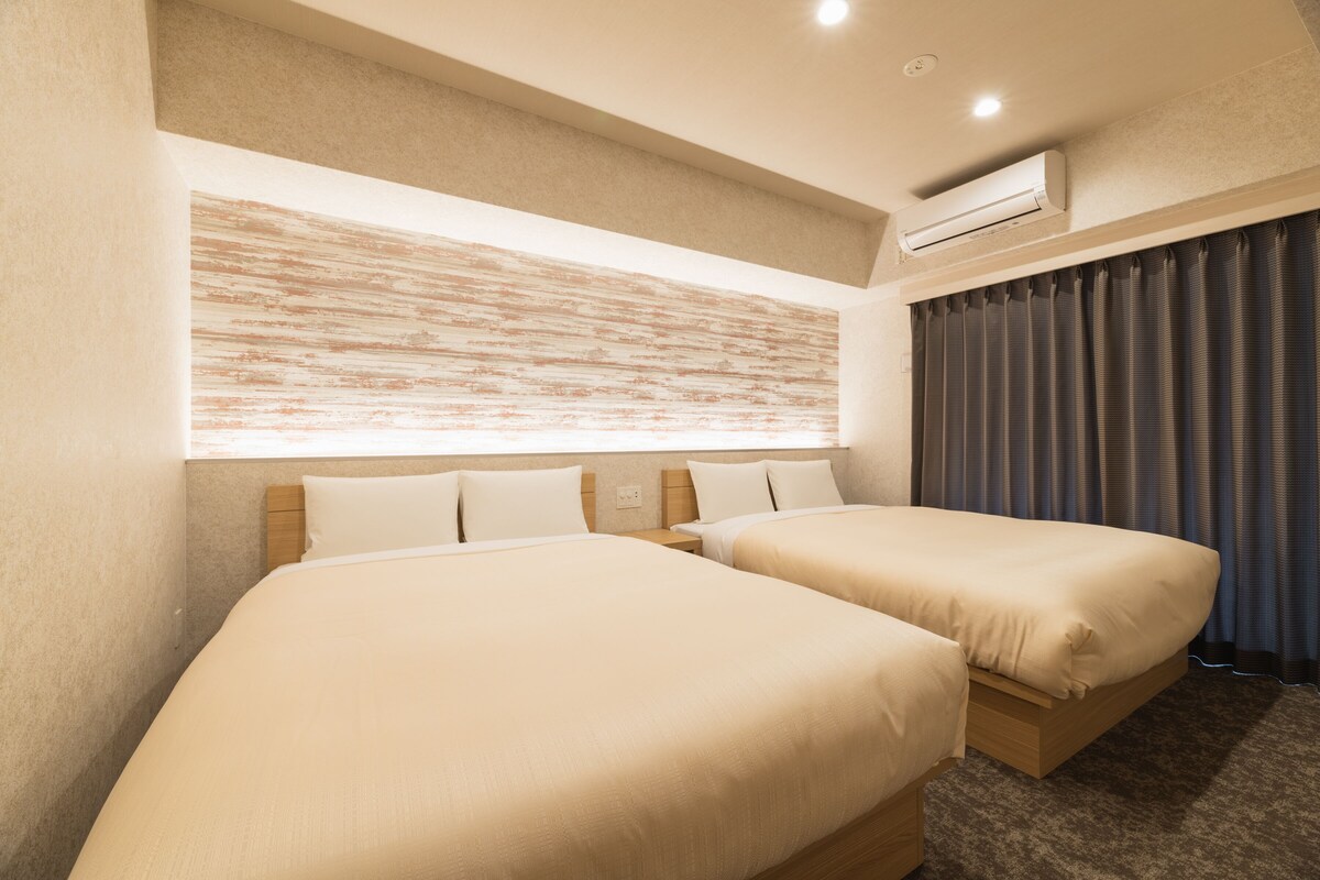 A hotel room with two beds, a large wall-mounted air conditioner, and a window with curtains.
