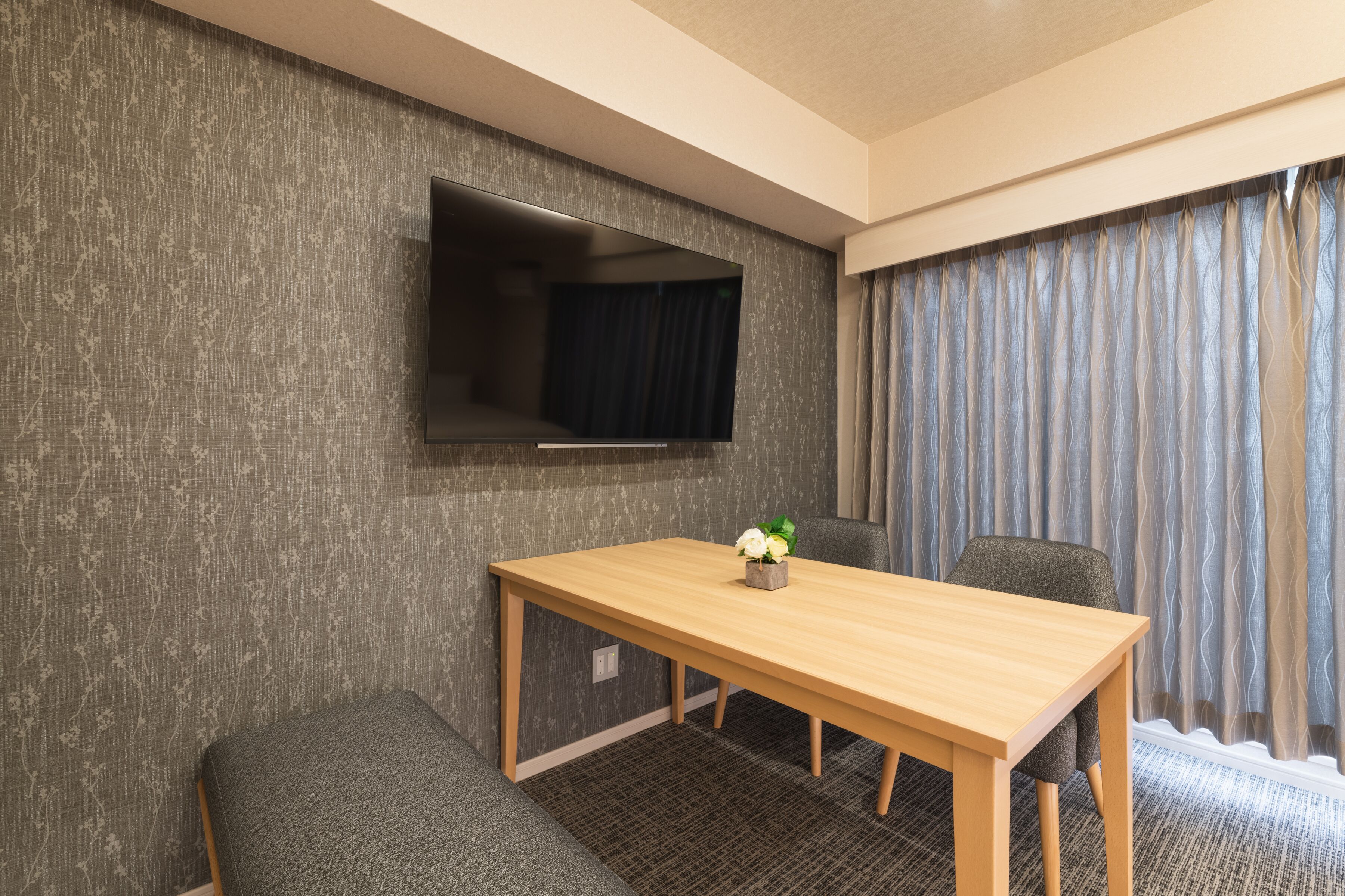 Habitación de hotel con una mesa de madera, dos sillones grises tapizados, un televisor de pantalla plana montado en la pared y una ventana con cortinas grises.