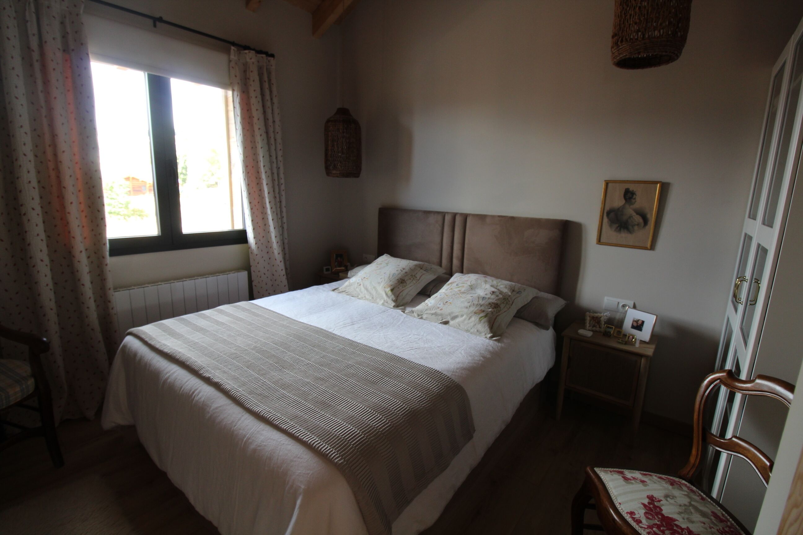 A bedroom with a bed, a chair, a nightstand, a window with curtains, a wall hanging, and a door.