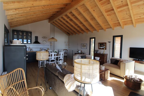 Un vaste espace de vie doté d’un haut plafond en bois, d’un canapé, d’une table à manger avec des chaises, et d’une cuisine à l’arrière-plan.