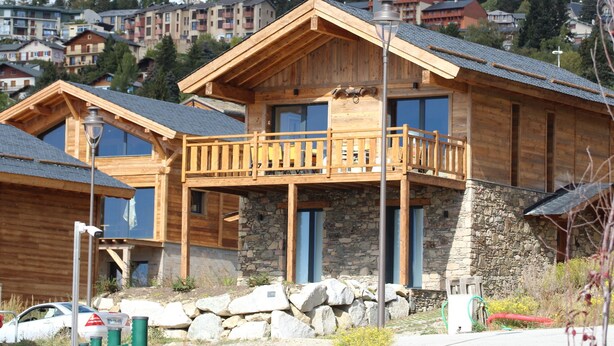 Una baita in legno con balcone, fondamenta in pietra e vista sugli edifici circostanti.