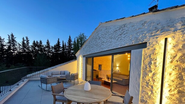 Una casa moderna con techo inclinado, un patio con una mesa redonda y sillas, y un cerramiento de vidrio que da a un espacio interior con un sofá y una lámpara.