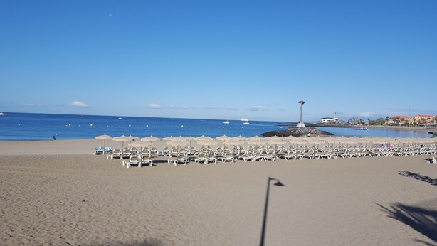 A beach with rows of sun loungers and umbrellas, a rocky outcrop with a monument, and boats on the water. - Best Location. Playa Las Vistas. Free Wifi (Playa de la Américas)