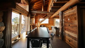 Interior de madera con una larga mesa de comedor y sillas, ventanas grandes y una escalera que lleva a un balcón.