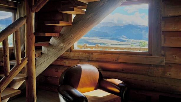 Interior de una cabaña de madera con escalera de madera, una silla y una ventana con vista a las montañas.