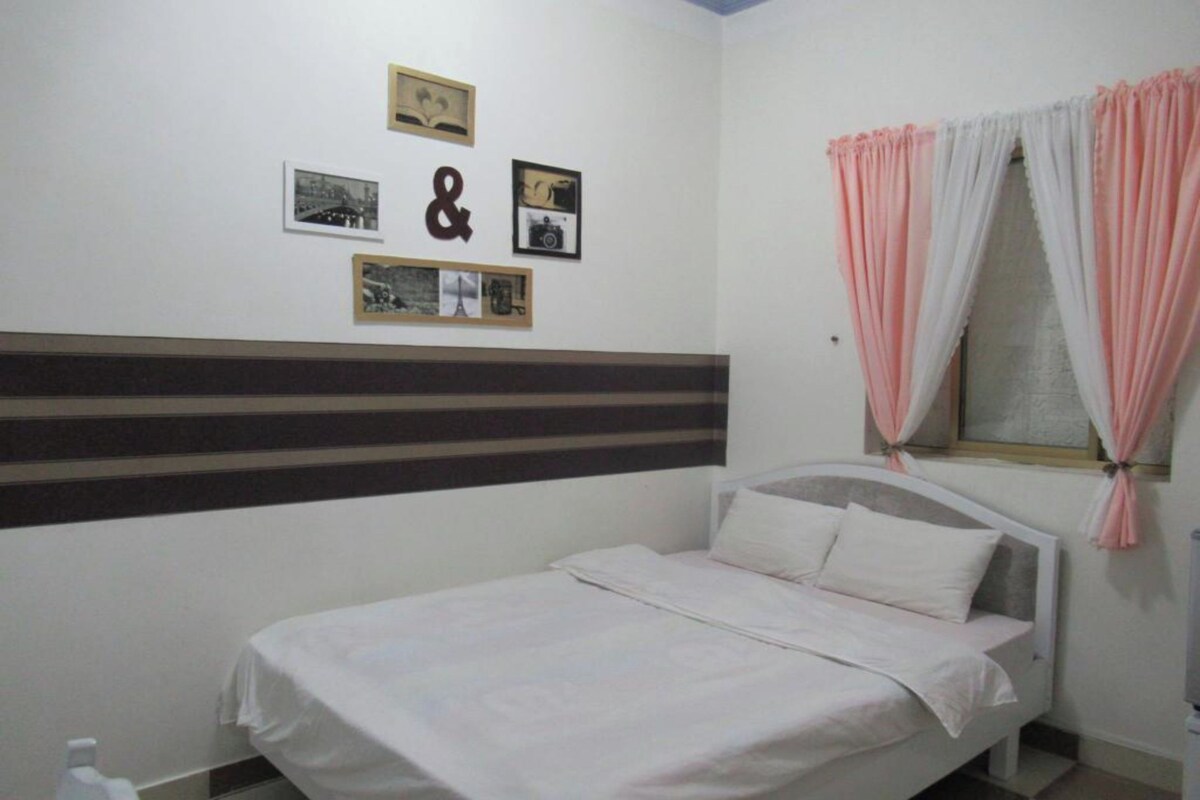 A single bed with a striped headboard, a window with pink and white curtains, and several framed pictures on the wall.