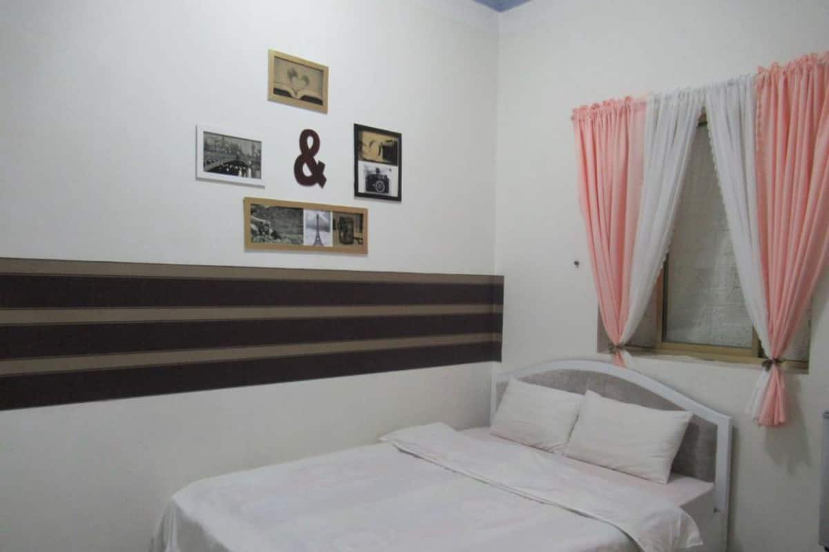 A bedroom with a bed, a window with pink curtains, and three framed pictures on the wall.