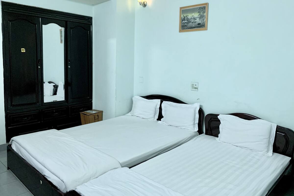 A hotel room with two single beds, a large wardrobe, a small bedside table, and a wall-mounted light fixture.