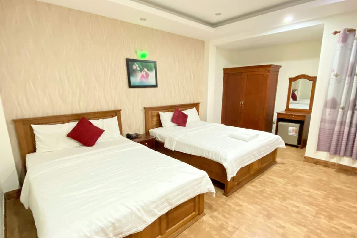 Une chambre d’hôtel avec deux lits en bois, du linge de lit blanc et des oreillers rouges. On y trouve une petite table de chevet avec un téléphone et une lampe verte au-dessus du lit. Une armoire en bois et un miroir sont visibles à l’arrière-plan.