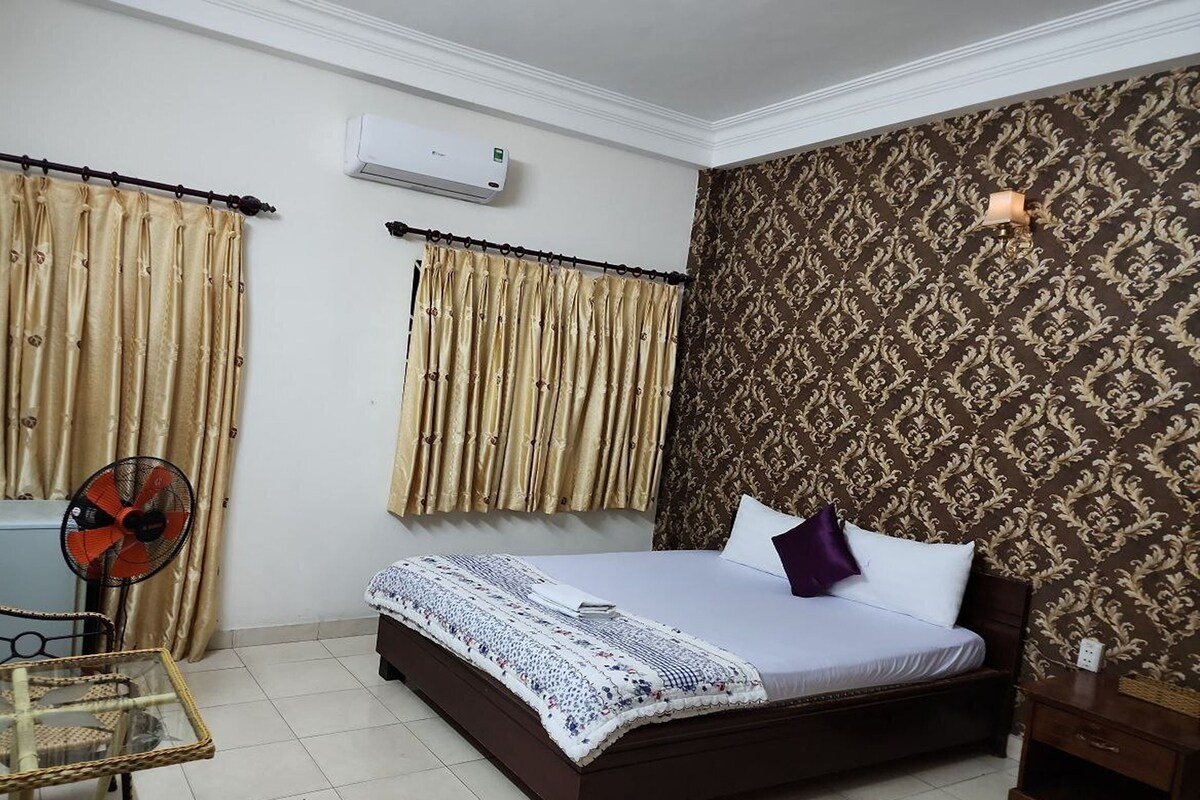 A hotel room with a bed, curtains, an air conditioning unit, a fan, a small table, and a patterned wall.