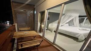 Varanda com mesa e cadeiras de madeira, porta de vidro deslizante que dá para um quarto com cama e dossel.