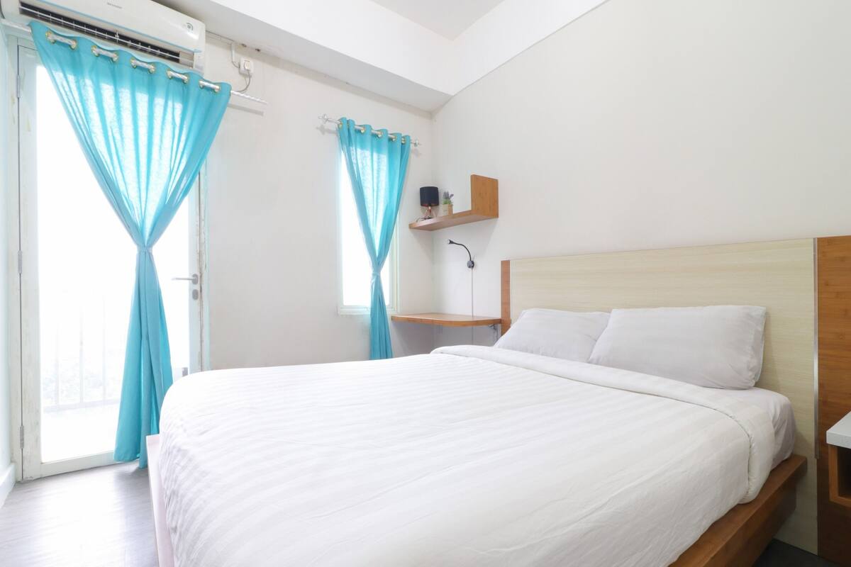 A bedroom with a bed, a nightstand, a shelf with a lamp, and a window with curtains.