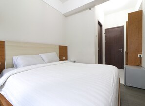 Una habitación de hotel con cabecera de madera, cama con ropa blanca, mesita de noche pequeña, armario y un calentador fijado a la pared.