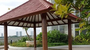 Una pérgola con piso de baldosas y techo rojo, rodeada de follaje y con un paisaje urbano al fondo.