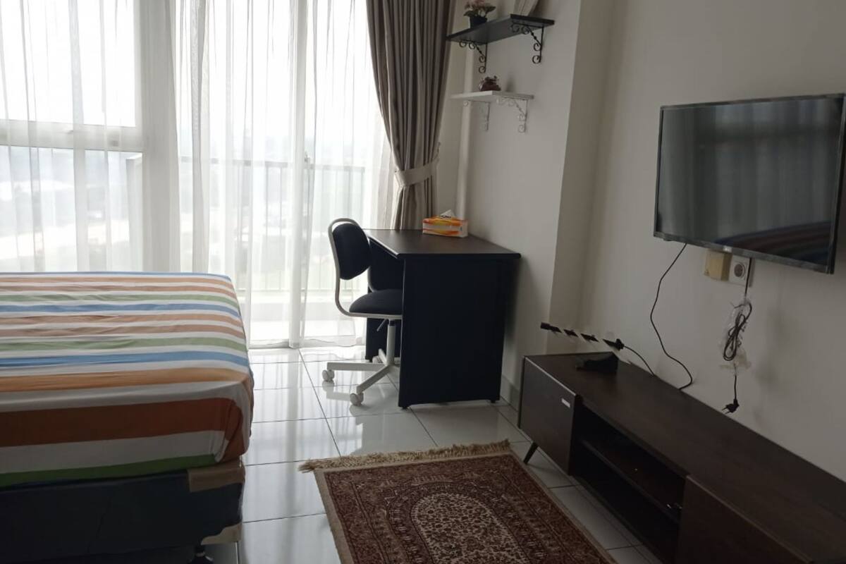Una habitación de hotel con cama, escritorio, silla, televisión y una pequeña estantería.
