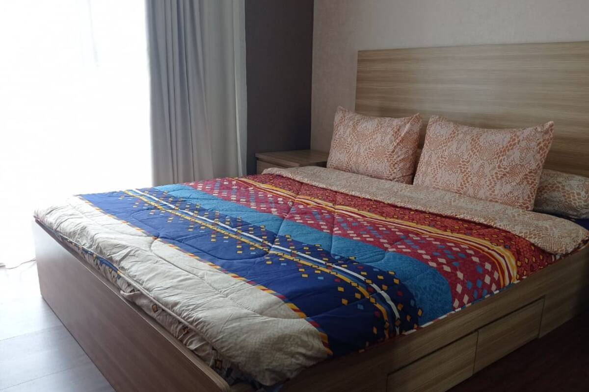 A bed with a patterned bedspread and two pillows, a wooden headboard, and a nightstand with a lamp.