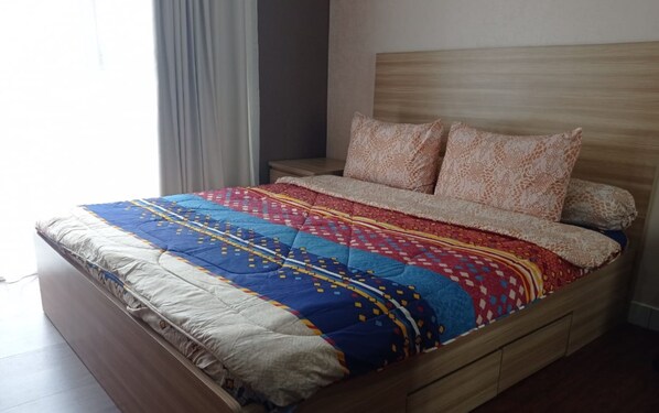 A bed with a patterned bedspread and two pillows, a wooden headboard, and a nightstand with a lamp. - Sultan Premiere Casa De Parco (Pagedangan)